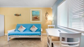 Ein Zimmer mit einem blauen Sofa, weißen Stühlen, einem kleinen Tisch, einer Lampe und einem Fenster mit Jalousien.