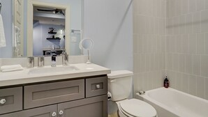 Une salle de bain avec un lavabo blanc, un miroir et des toilettes. On y trouve un porte-serviettes et une bouteille de shampoing posée sur le lavabo.