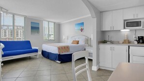 A modern hotel room with a bed, a blue sofa, a kitchenette, and large windows with city views. - Pelican Pointe 317:Walk to Beach~Pool~Free Parking (Clearwater Beach)