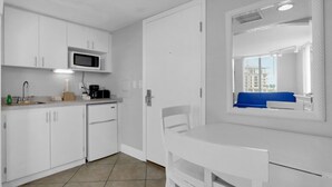 A compact kitchenette with a microwave, coffee maker, and a small refrigerator. There is a window with a view of buildings and a blue chair. - Pelican Pointe 317:Walk to Beach~Pool~Free Parking (Clearwater Beach)