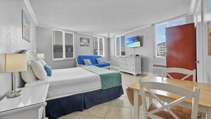 A bedroom with a bed, bedside table, lamp, dresser, TV, and a dining area with a table and chairs. - Pelican Pointe 318:Walk to Beach~Pool~Free Parking (Clearwater Beach)
