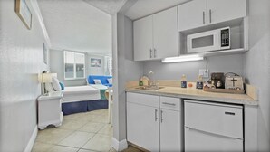 A compact hotel room with a kitchenette, a bed with blue bedding, a bedside table with a lamp, and a view of the outside through a window. - Pelican Pointe 318:Walk to Beach~Pool~Free Parking (Clearwater Beach)