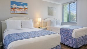 A hotel room with two beds, a nightstand with a lamp, a painting of beach umbrellas, and a window with blinds.