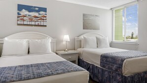 Ein Hotelzimmer mit zwei Betten, einem Nachttisch mit Lampe, einem Bild von Strandsonnenschirmen und einem Fenster mit Jalousien.