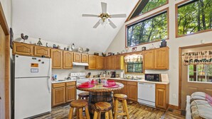 Une cuisine avec des meubles de rangement en bois, un îlot central doté de tabourets, un ventilateur de plafond et une grande fenêtre donnant sur la forêt.