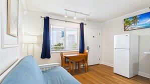 Una habitación compacta con sofá, una mesa de comedor con sillas, un refrigerador blanco y una ventana con cortinas.