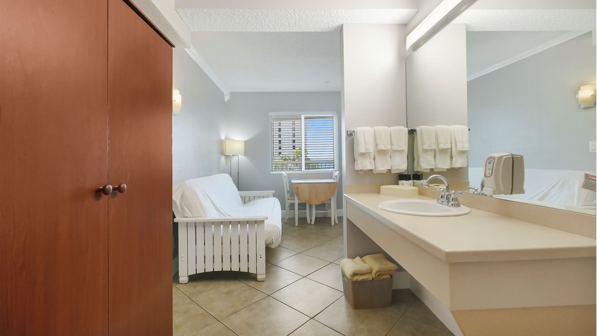 Un baño moderno con un lavabo blanco, espejo y perchero. Se observa una cuna blanca con cambiador y una ventana con persianas.