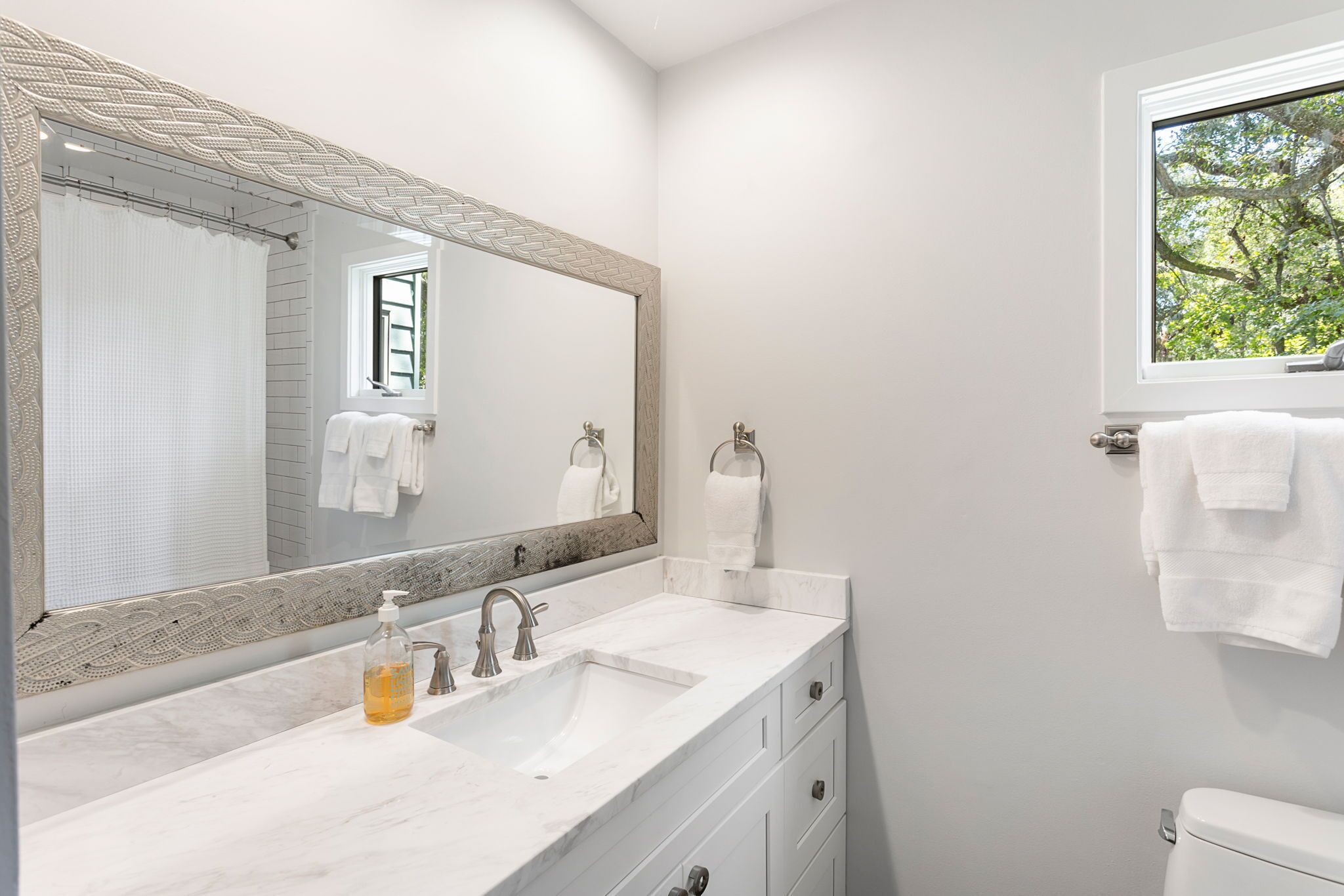 A modern bathroom with a large mirror, white fixtures, and a window providing a view of trees.