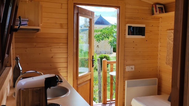 A wooden cabin interior with a kitchen area, a bed, and a view of a stone building outside.