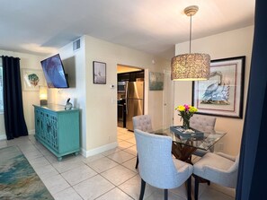 A dining area with a glass table, chairs, a teal cabinet, a hanging light fixture, and a framed picture on the wall.