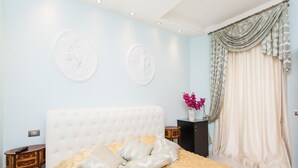 A bedroom with a white tufted headboard, a gold patterned bedspread, two wall-mounted decorative plates, a bedside table with a vase of flowers, and a large window with sheer curtains. - Superior Suite (St. Peter Vatican Bambino Gesù Gianicolo) (ROMA)