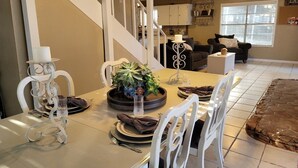 Comedor con una mesa dispuesta para cuatro personas, sillas blancas, un centro de mesa decorativo y una escalera que sube a un nivel superior.