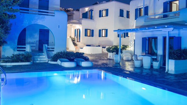 Ein Poolbereich mit einem klaren, blauen Pool, weißen Gebäuden mit blauen Fensterläden und Außenmöbeln.