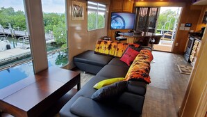 A modern living area with a large window overlooking a dock and water, a black sofa with colorful cushions, a wooden table, and a kitchen area with a TV. - Key West Luxury Houseboat (Key West)