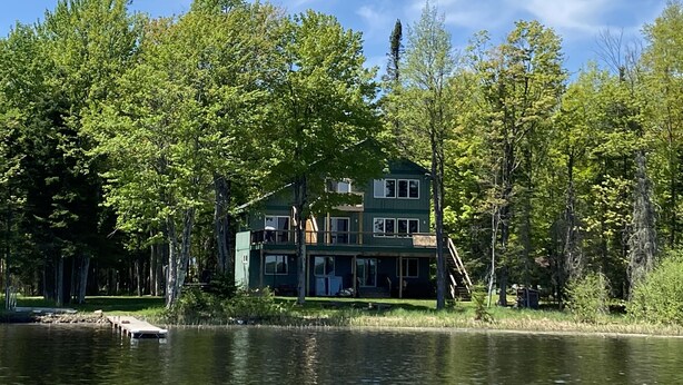 A two-story house with a dock extending into a lake, surrounded by dense trees.