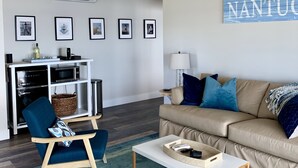 Ein modernes Wohnzimmer mit einem beige farbenen Sofa, blauen Sesseln, einem Couchtisch und einer Deckenleuchte. An den Wänden befinden sich gerahmte Bilder und ein Schild mit der Aufschrift 'NANTU'.