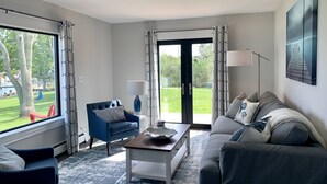 A modern living room with a sofa, armchair, coffee table, and a view of a garden through large windows. - Harbor View Landing Mystic, CT - The Bluffs Waterfront House - First Floor ONLY (Mystic)