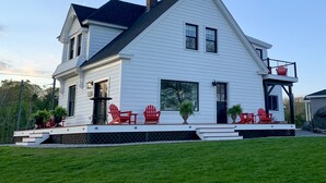 A two-story white house with a dark roof, a front porch with red Adirondack chairs, and a small deck area. - Harbor View Landing Mystic, CT - The Bluffs Waterfront House - First Floor ONLY (Mystic)