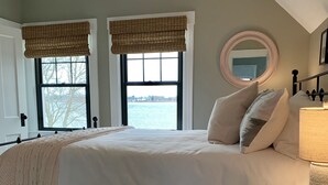A bedroom with a large bed, two windows with blinds, a round mirror, and a bedside table with a lamp.