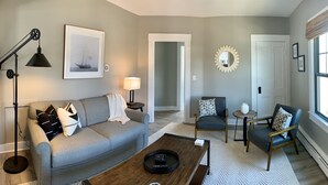 A living room with a grey sofa, a coffee table, a floor lamp, a side table with a lamp, a chair, and a window with blinds.