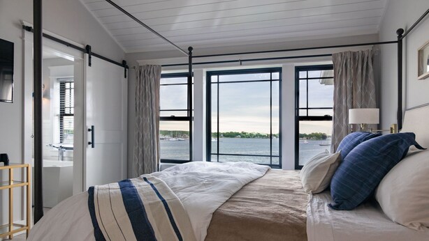 A bedroom with a large window offering a view of the water and boats. There is a bed with a striped blanket and pillows, a bedside table, and a lamp.