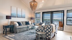 A modern living room with a sofa, coffee table, and patterned armchair. Large windows offer a view of the water.