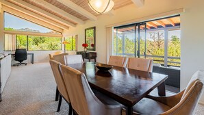 Un comedor amplio con una gran mesa de madera y sillas cómodas, una lámpara moderna y ventanas grandes que ofrecen vistas a la vegetación y montañas.