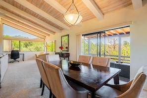 Un comedor amplio con una gran mesa de madera y sillas cómodas, una lámpara moderna y ventanas grandes que ofrecen vistas a la vegetación y montañas.