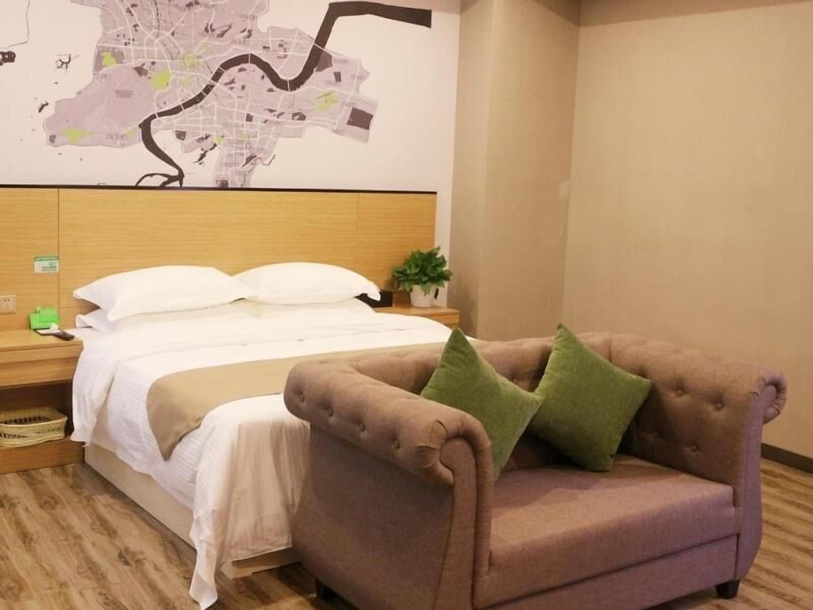 A hotel room with a bed, a map on the wall, a wooden headboard, a nightstand, a green plant, and a brown armchair.