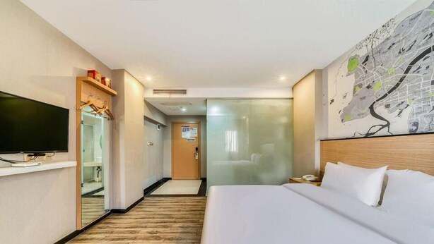 A hotel room with a large bed, a flat-screen TV, a wooden floor, a map on the wall, and a hallway leading to another room.