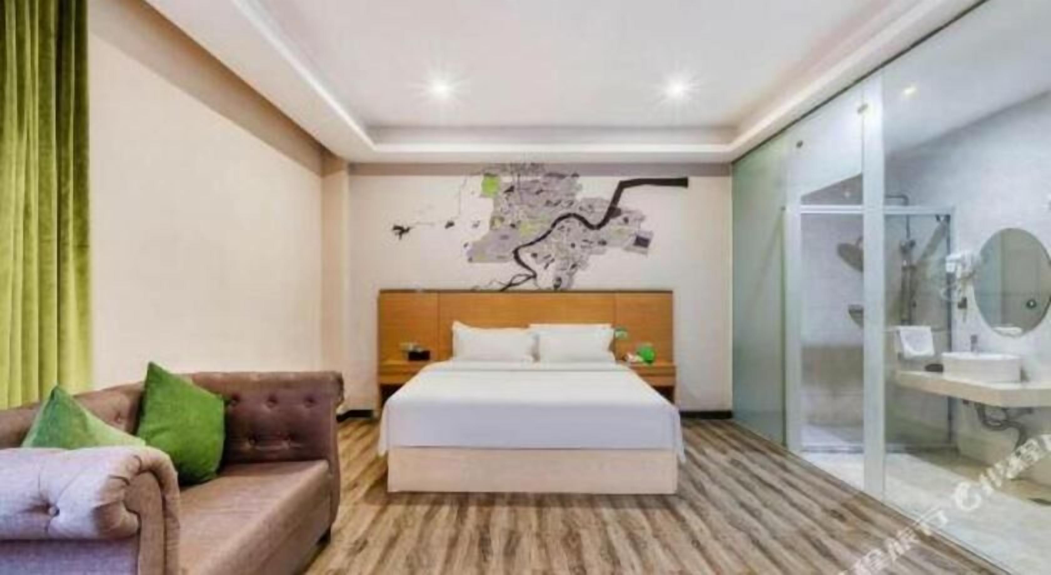 A hotel room with a bed, a wooden headboard, a map artwork on the wall, a green cushion on a sofa, and a bathroom with a mirror and sink.