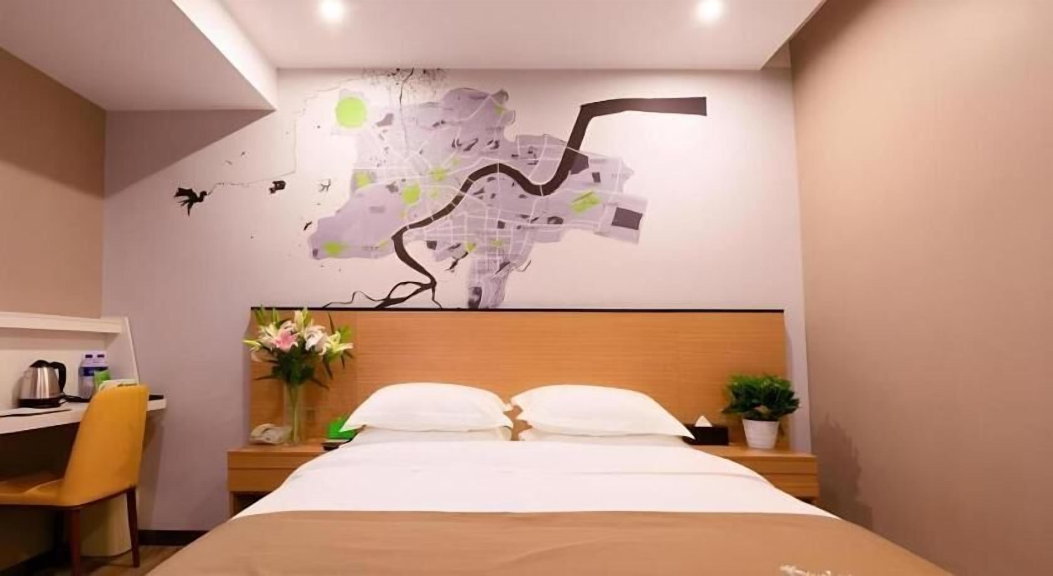 A hotel room with a bed, a desk, a chair, a nightstand with a vase of flowers, and a wall with a map design.