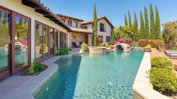 Una casa moderna con piscina, muros de piedra y un jardín con árboles altos y arbustos.