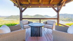 Una terraza de madera con mobiliario de mimbre y una mesa redonda, con vistas a un valle y colinas.