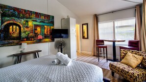 Habitación de hotel con cama, un cuadro, televisor, ventilador, mesa y vista al exterior por la ventana.
