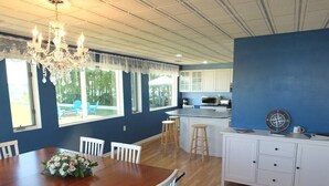 Un coin repas avec une table en bois, des chaises blanches et un lustre. On y trouve également une cuisine avec des meubles blancs et un mur d’accent bleu.