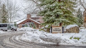 Entrada de um chalé coberto de neve com uma placa indicando "Sunriver Lodge", um edifício de madeira e um ônibus estacionado.