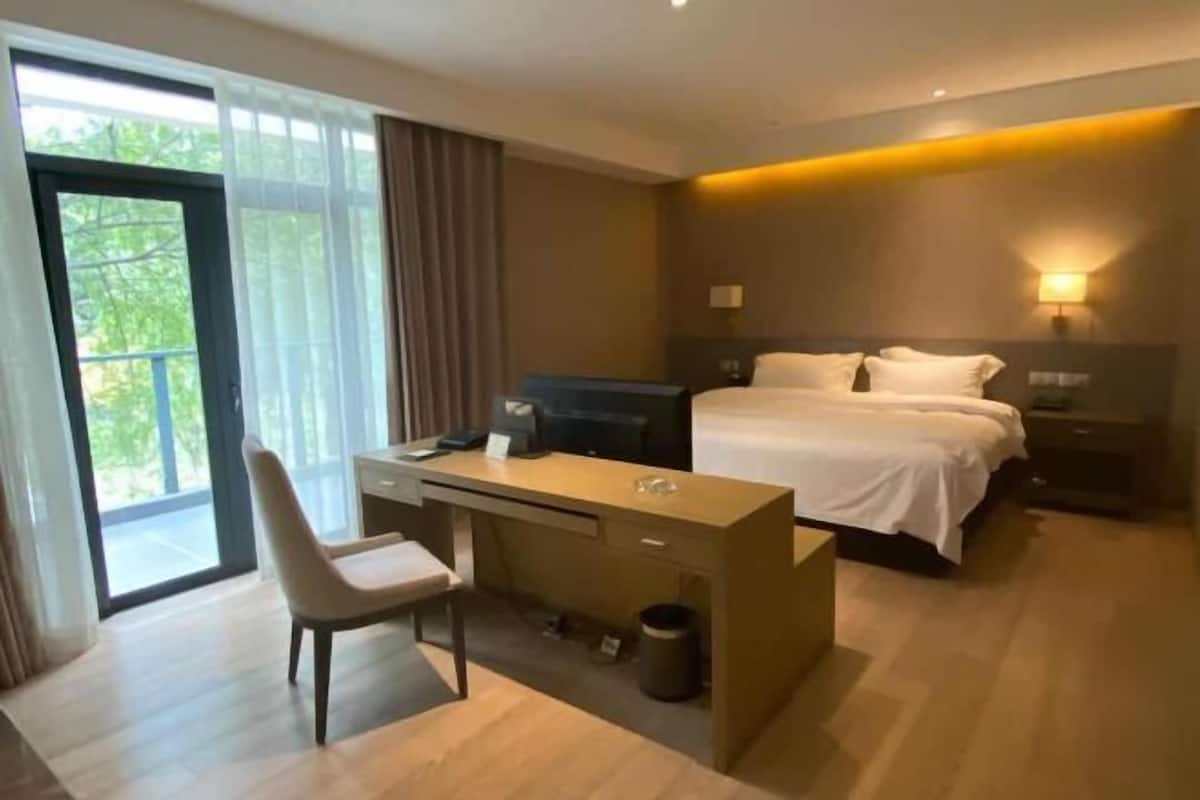 A hotel room with a bed, desk, chair, and a view of greenery through the window.