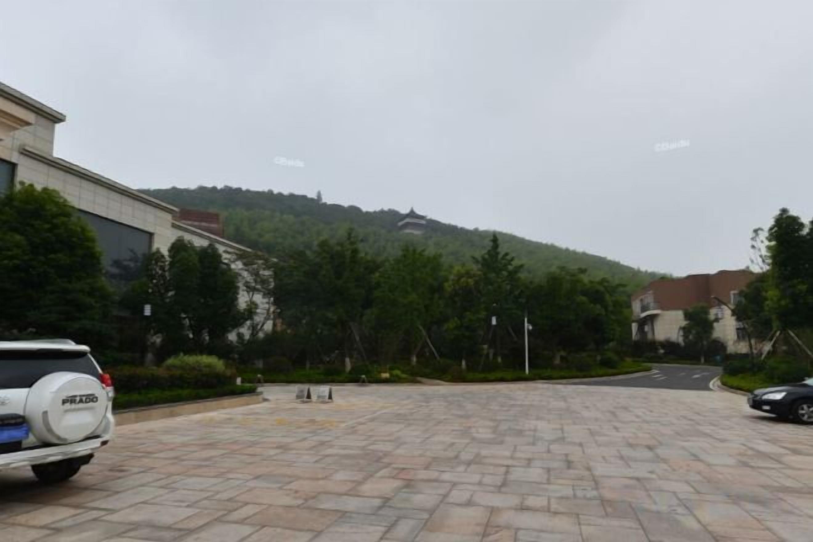 A paved parking area with a white SUV, buildings, and a hilly landscape in the background.