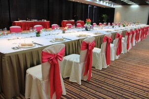 Un salón de banquetes con largas mesas dispuestas para un evento formal, cada una con mantel blanco y fundas rojas para las sillas.