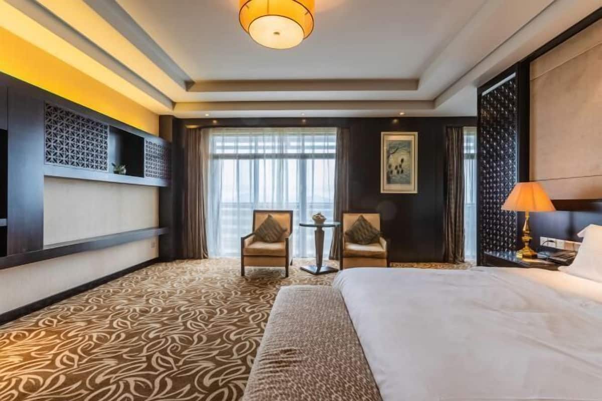 A hotel room with a large bed, two armchairs, a small table, a decorative wall, and a large window with curtains.