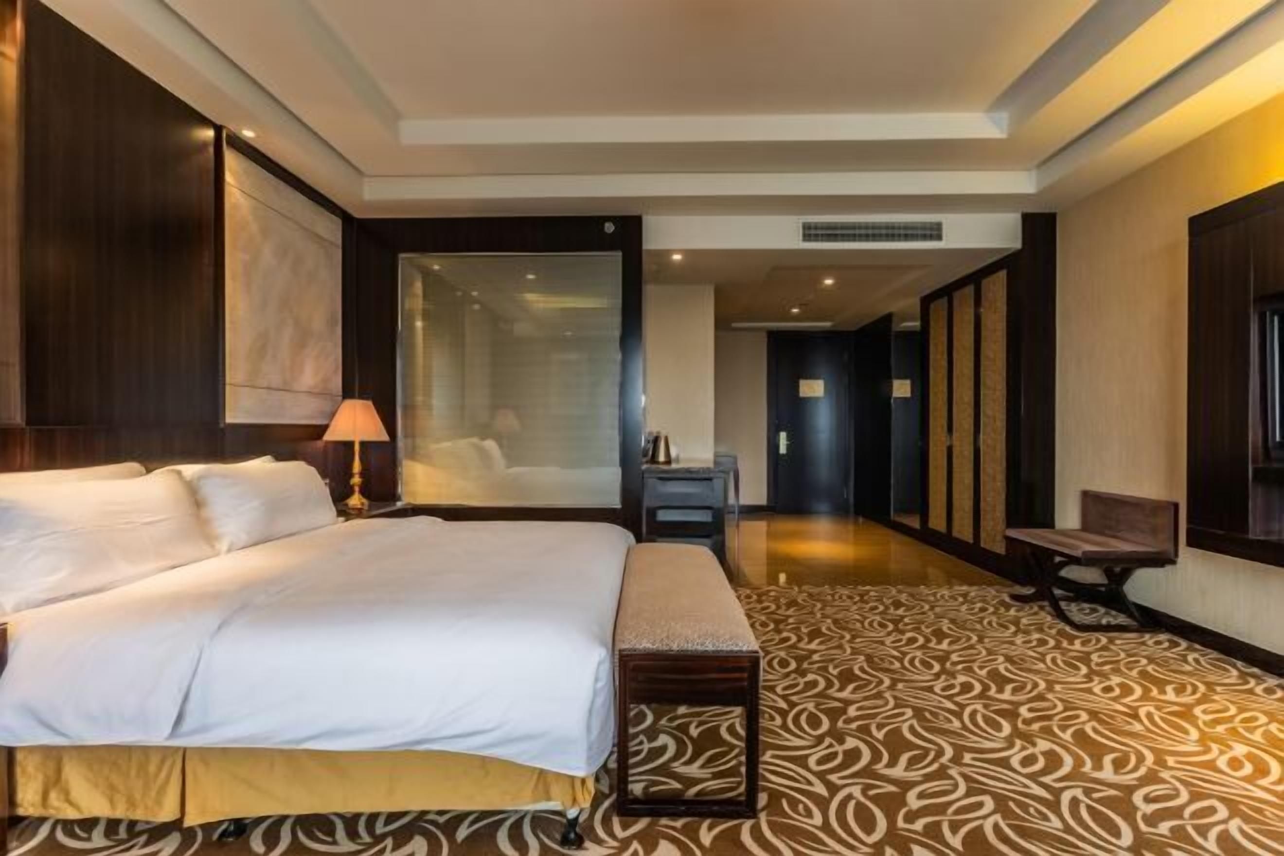 A hotel room with a large bed, a bedside table, a lamp, a bench, and a patterned carpet.
