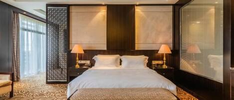 A hotel room with a large bed, two bedside lamps, a patterned carpet, and a large window with blinds.
