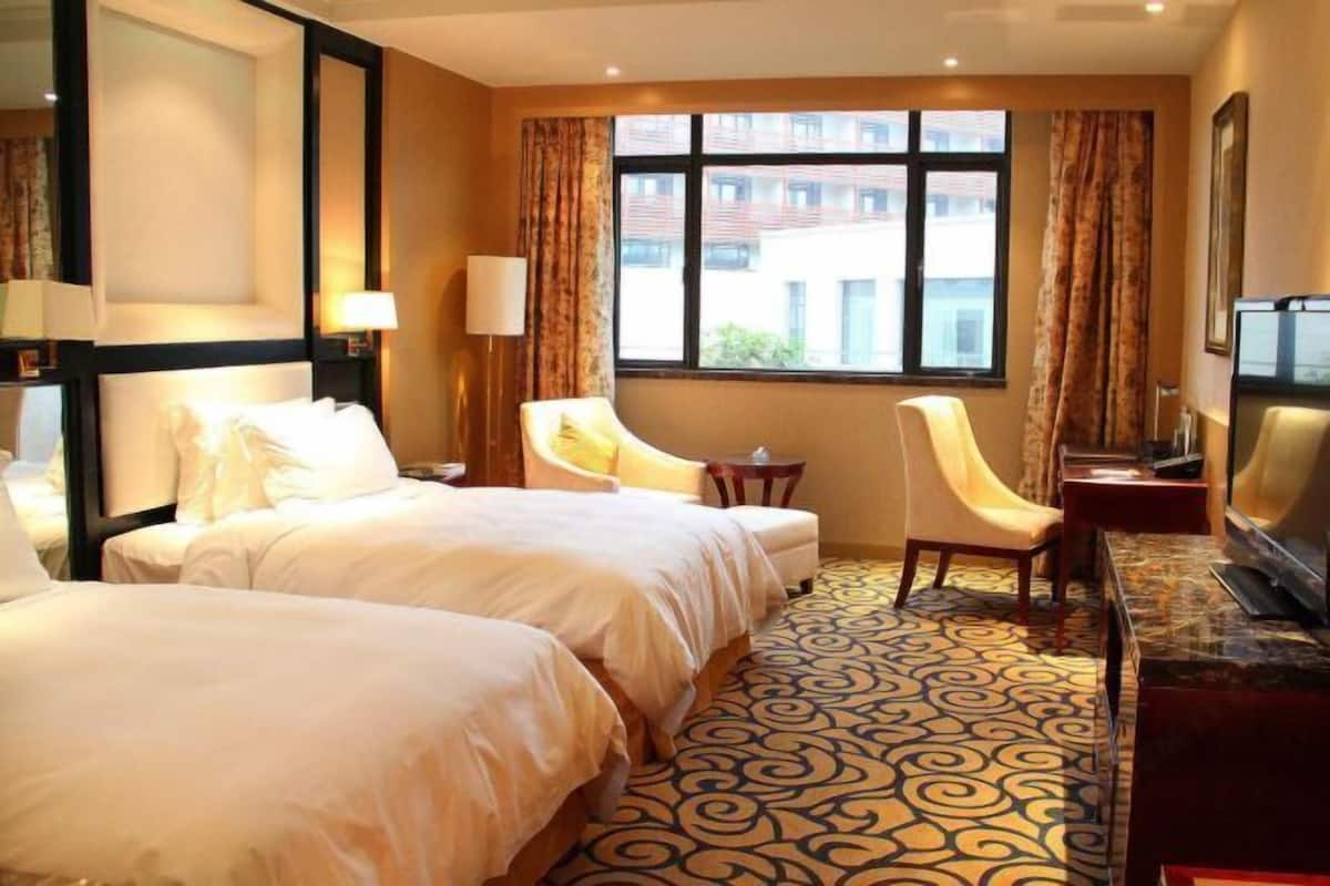 A hotel room with two beds, a desk with a chair, a television, and a large window with curtains.