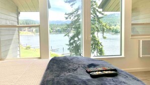 Ein Zimmer mit einem Bett, einer Fernbedienung und großen Fenstern mit Blick auf einen See und einen Wald.