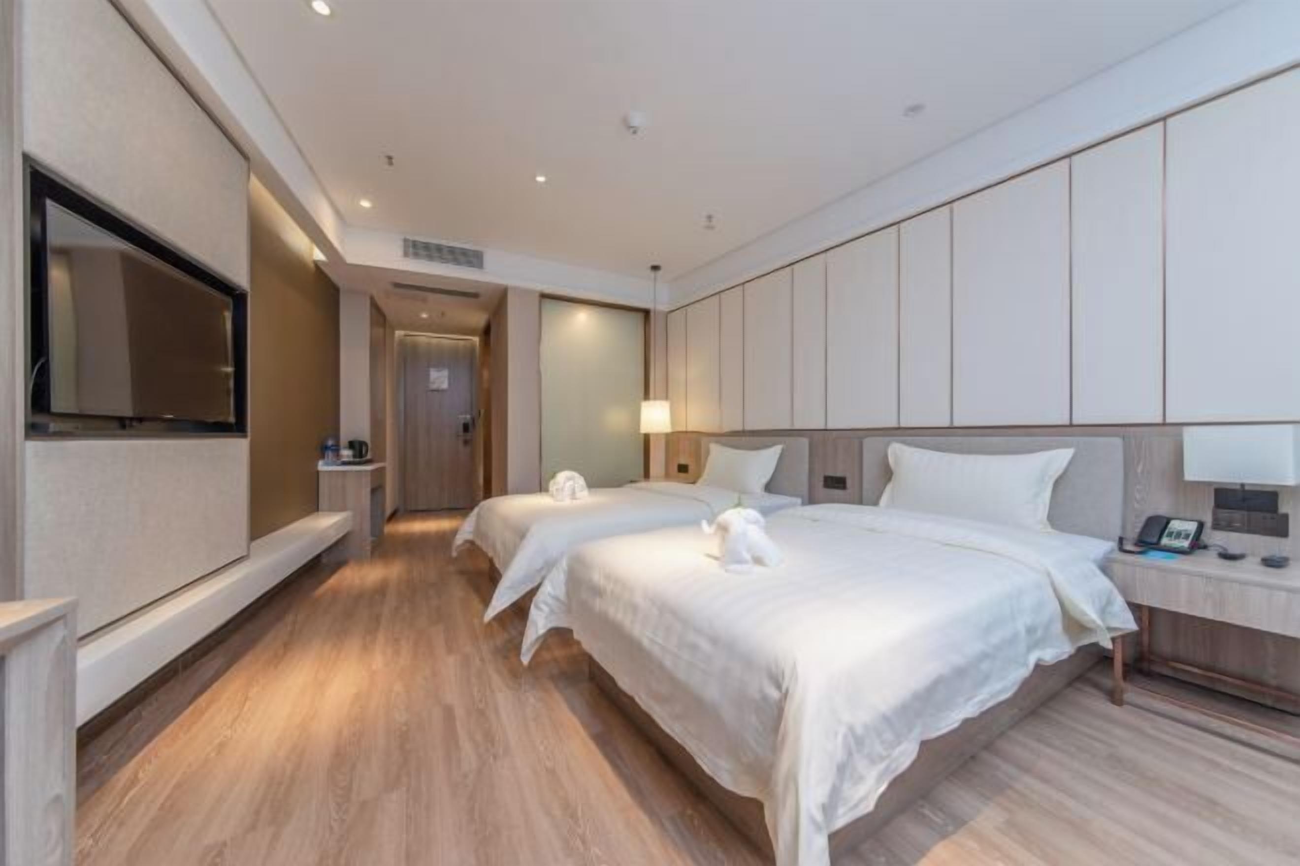 A hotel room with two beds, a TV, a bedside table with a lamp, and a wooden floor.