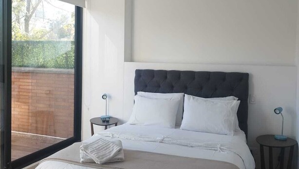 A modern bedroom with a large bed, a view through a glass door, and a small bedside table with a lamp. - Balmoral 103 (Bogotá)