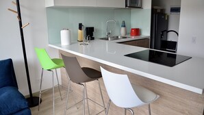 A modern kitchen with a white countertop, stainless steel sink, and a black refrigerator. There are three bar stools and a blue chair. - Balmoral 103 (Bogotá)