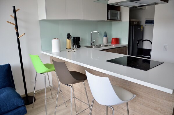 Une cuisine moderne avec un plan de travail blanc, un évier en acier inoxydable et un réfrigérateur noir. On y trouve trois tabourets de bar et une chaise bleue.