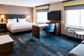 A hotel room with a bed, desk, chair, TV, and large windows.